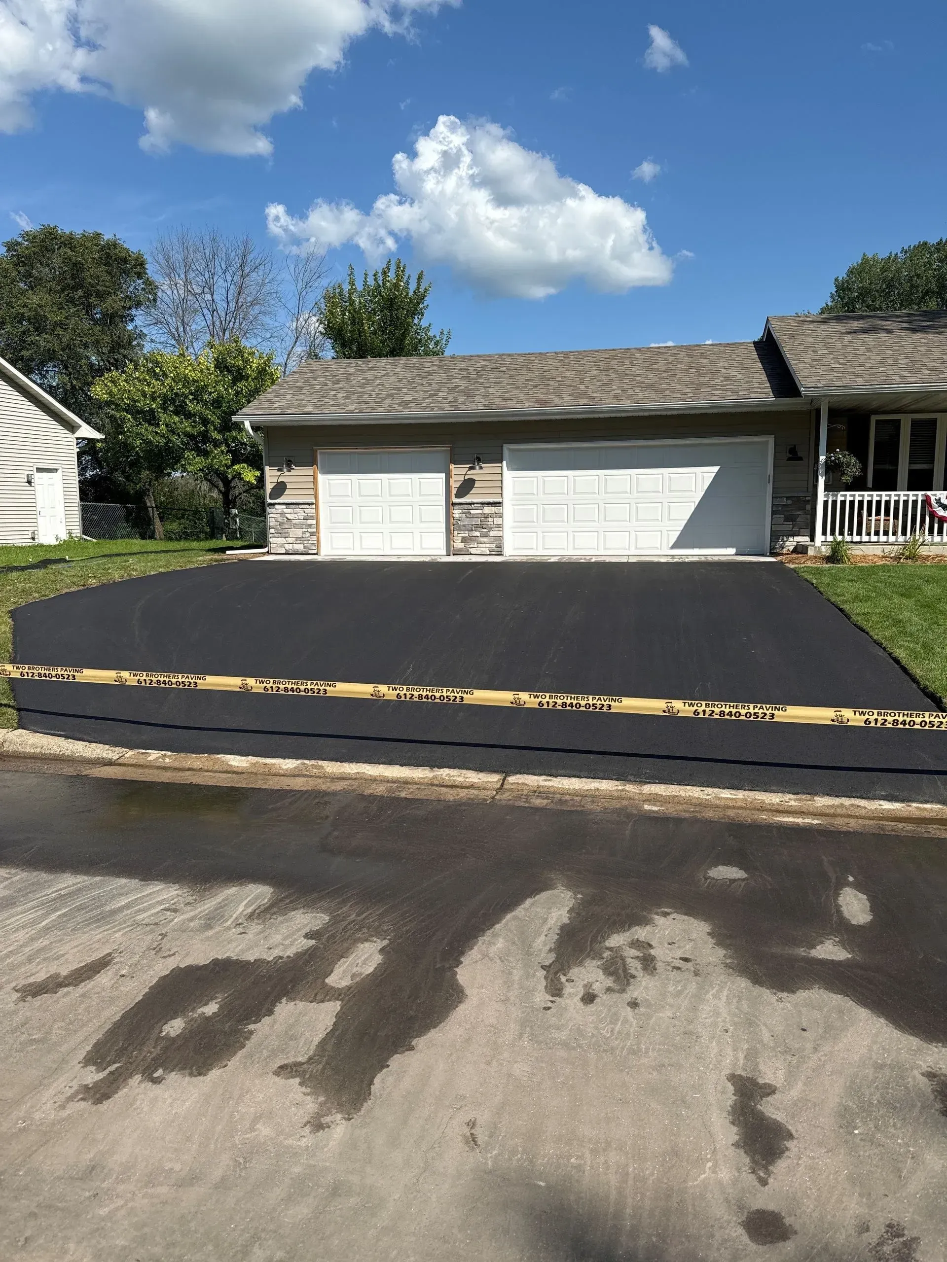 Freshly paved black driveway in front of a two-car garage on a sunny suburban street.