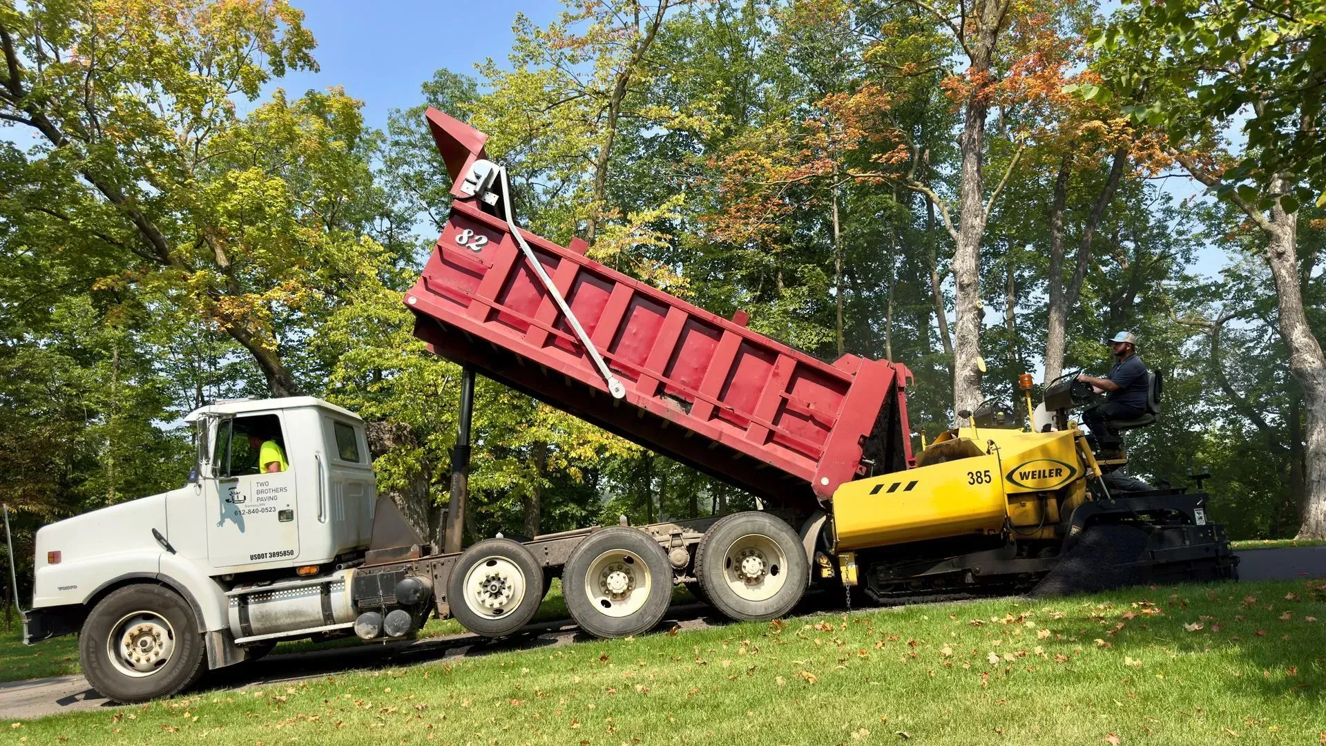 White dump truck with raised red bed and yellow asphalt paver on a grassy roadside with trees.