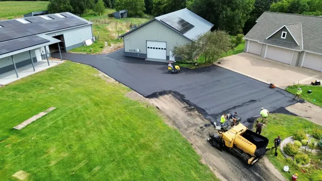 Workers paving a black driveway beside a house, garage, and yellow asphalt roller.