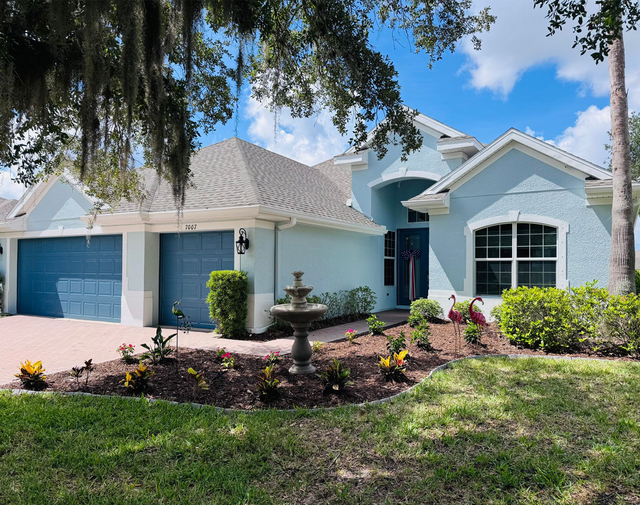 Single-story blue house with two-car garage, front fountain, and landscaped yard under a bright sky