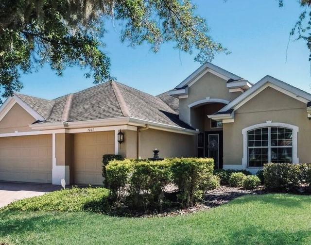 Suburban single-story house with beige exterior, white trim, and a manicured front lawn under a blue sky