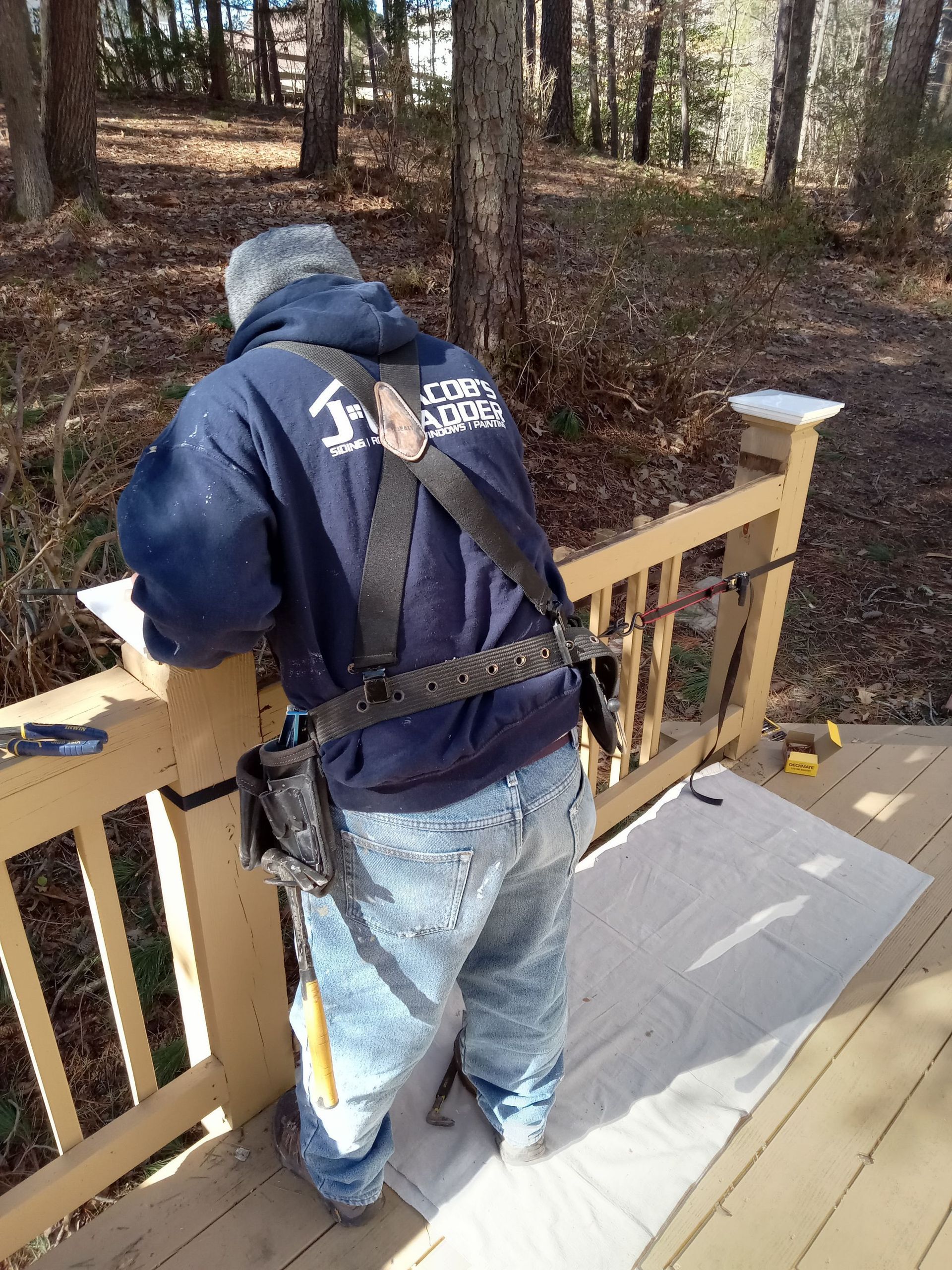Person in blue sweatshirt and jeans on a wooden deck, working. Safety harness, tools, and paint on deck.