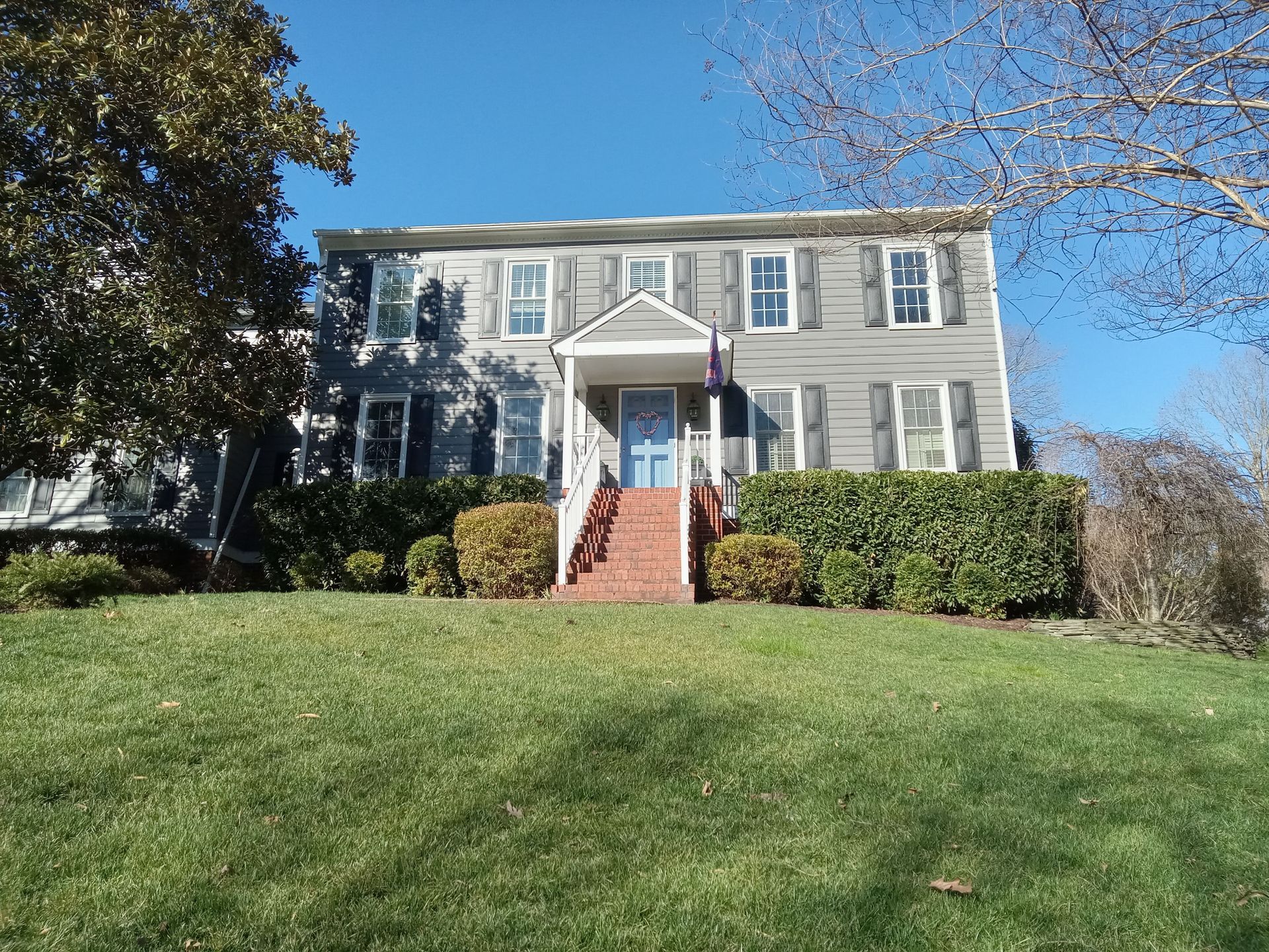 Two-story gray house with blue front door, white trim, and shutters; red brick stairs, green grass, and bushes.