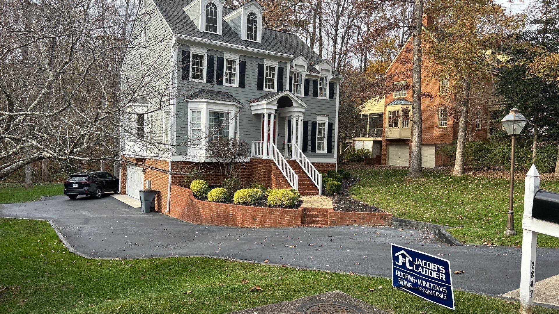 Gray house with black shutters, brick base, and a black car in driveway.