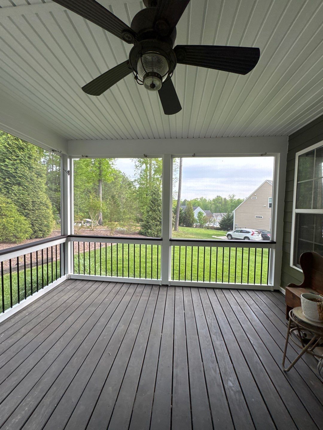 Screened-in porch with dark gray floorboards, black railing, and view of trees and houses. Ceiling fan overhead.