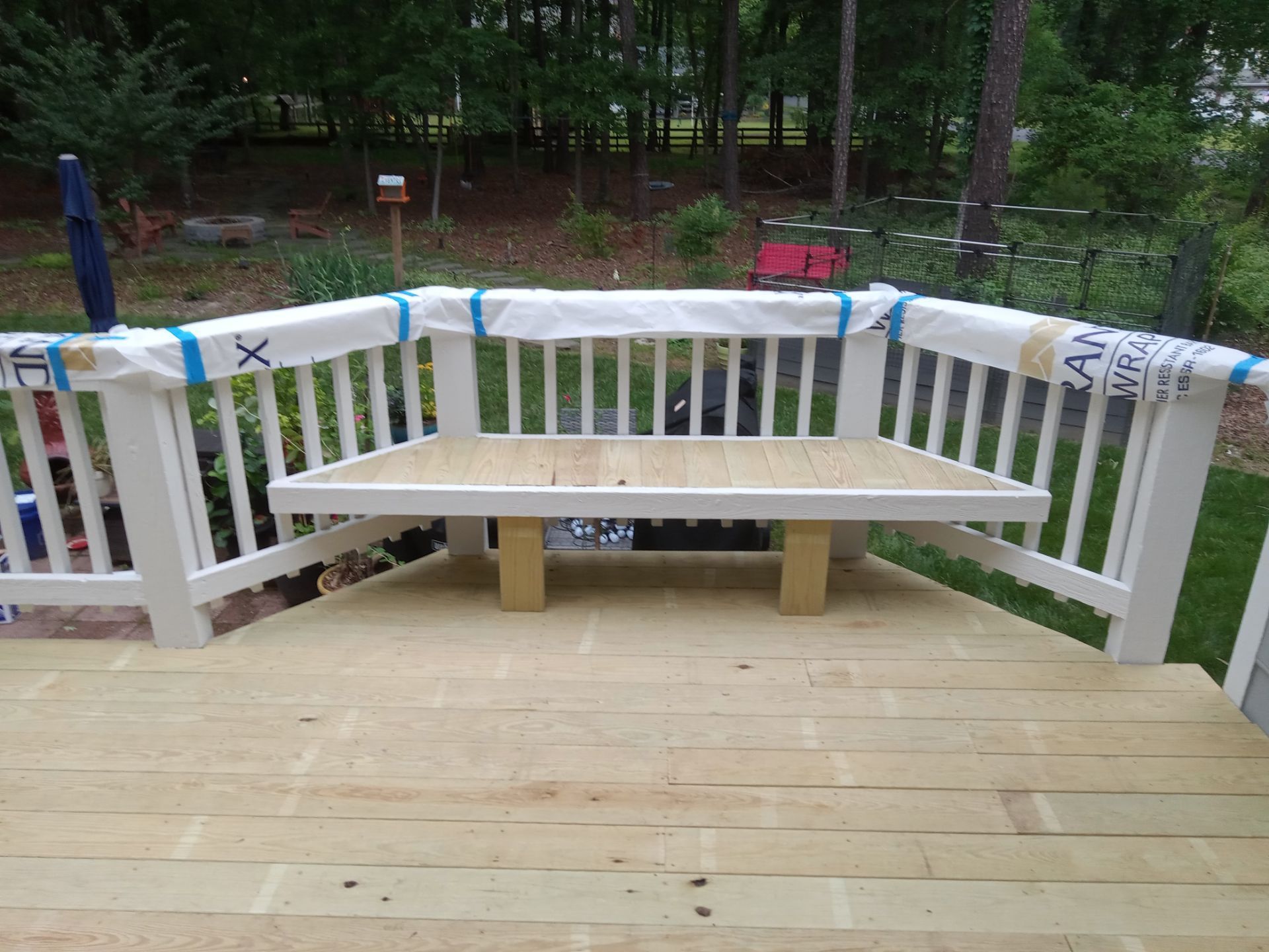 Deck with a built-in bench. White railing and natural wood deck and bench. Green trees in the background.