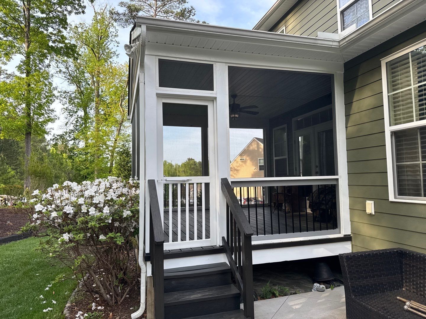 Screened porch with white trim, dark steps, and railing. Green siding of a house, landscaping, and trees in background.