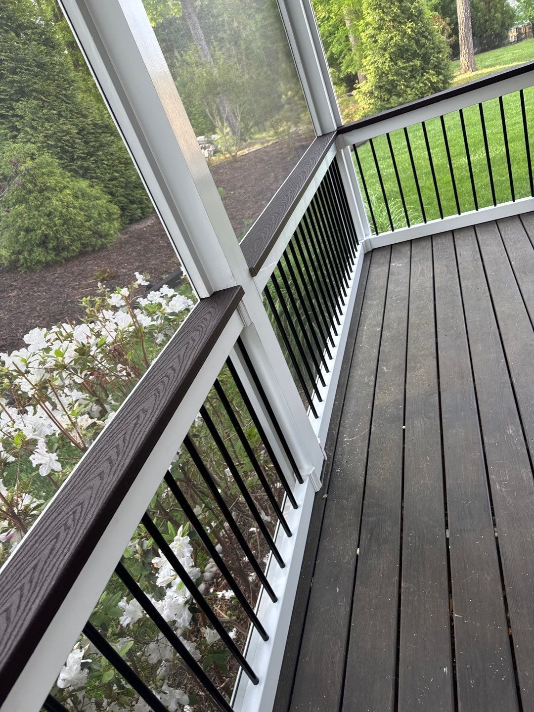 Screened porch corner with dark wood deck, black railing, white posts, and green yard in background.