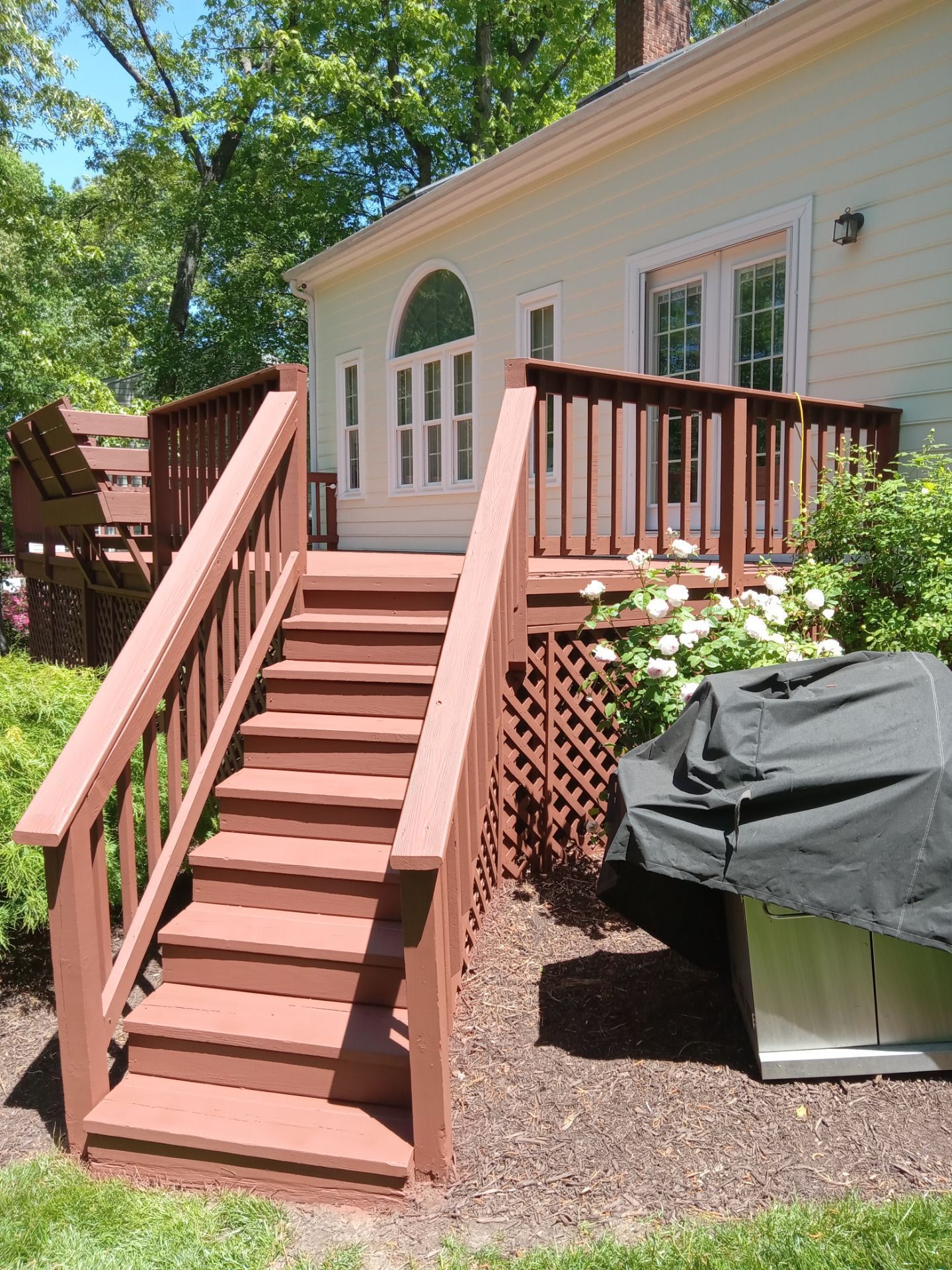 Wooden deck with stairs leading to a light yellow house with arched windows and a covered grill.