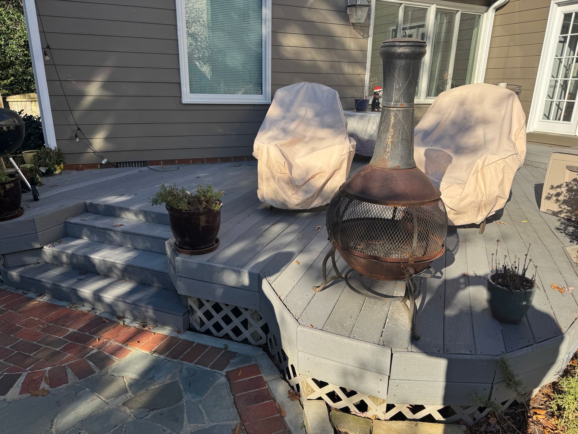 Wooden deck with fire pit, covered chairs, steps, and potted plants.