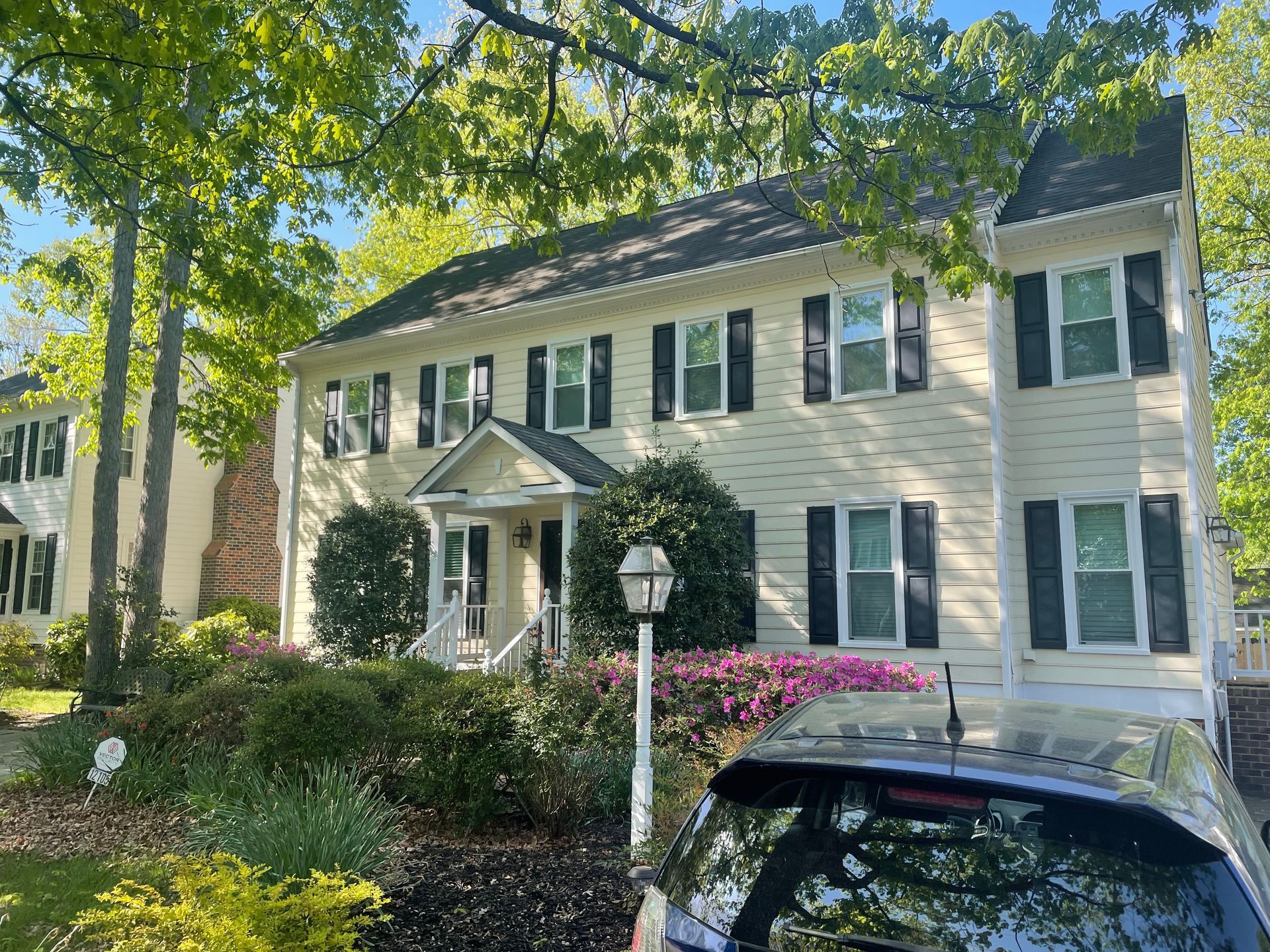 Two-story beige house with black shutters, white trim, and flowering bushes, car parked in front.