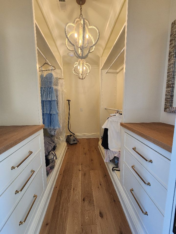 Narrow walk-in closet with white cabinets, wood floors, and two ornate chandeliers.