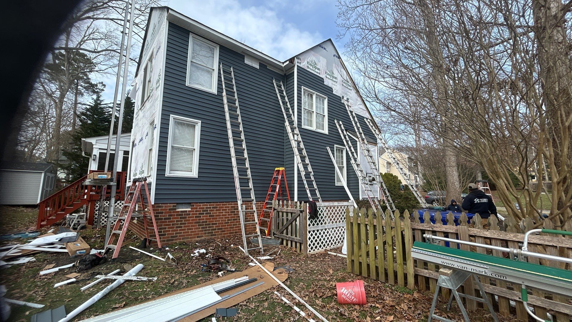 House exterior with dark blue siding replacement in progress; ladders, materials, and workers present.