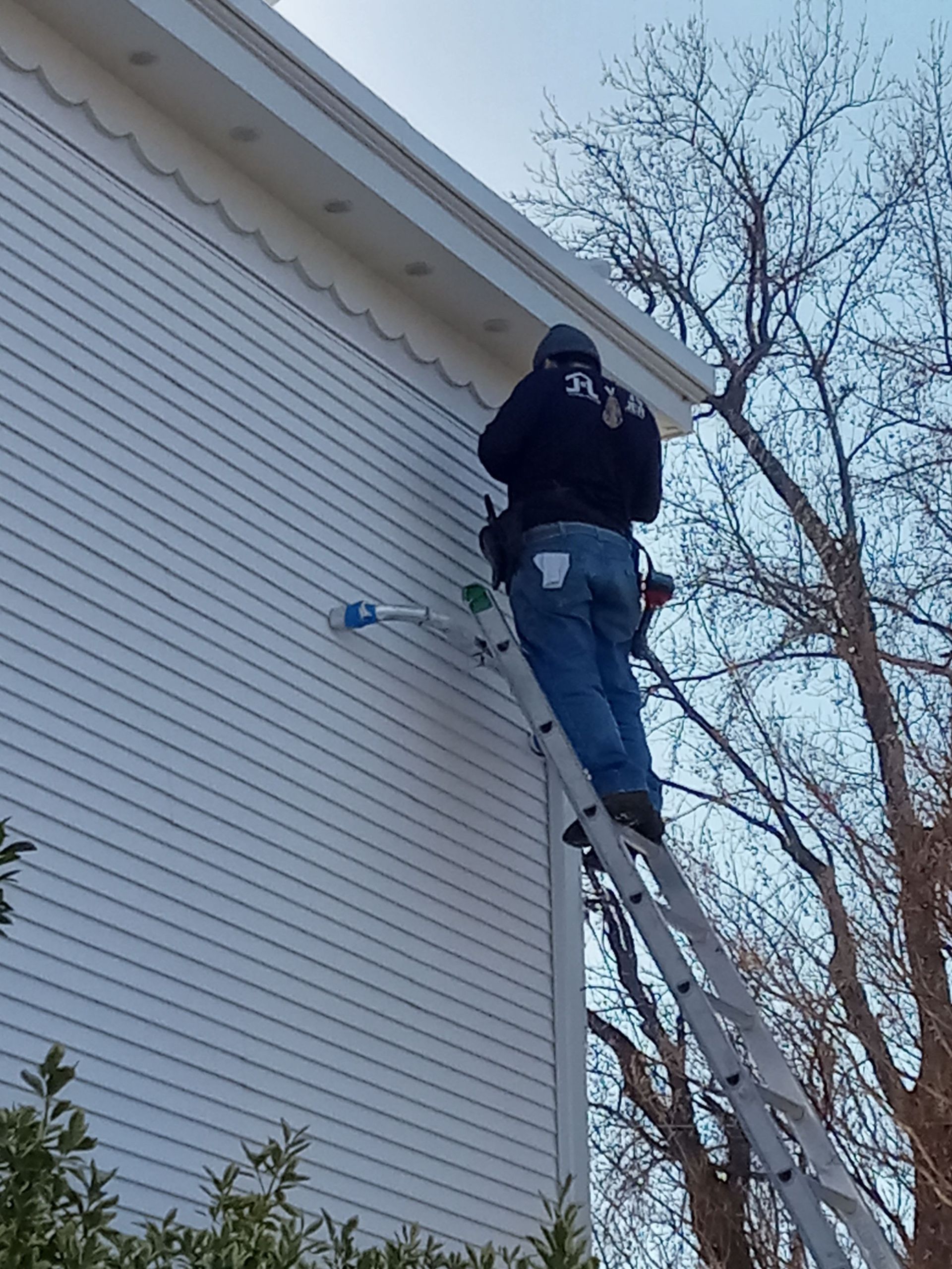 Person on ladder next to white house, working near the roofline, with leafless tree in the background.