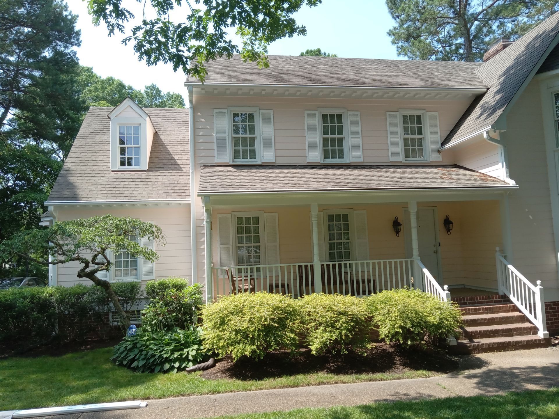 Two-story beige house with white shutters, porch, and dormer. Green shrubs and trees surround the house.