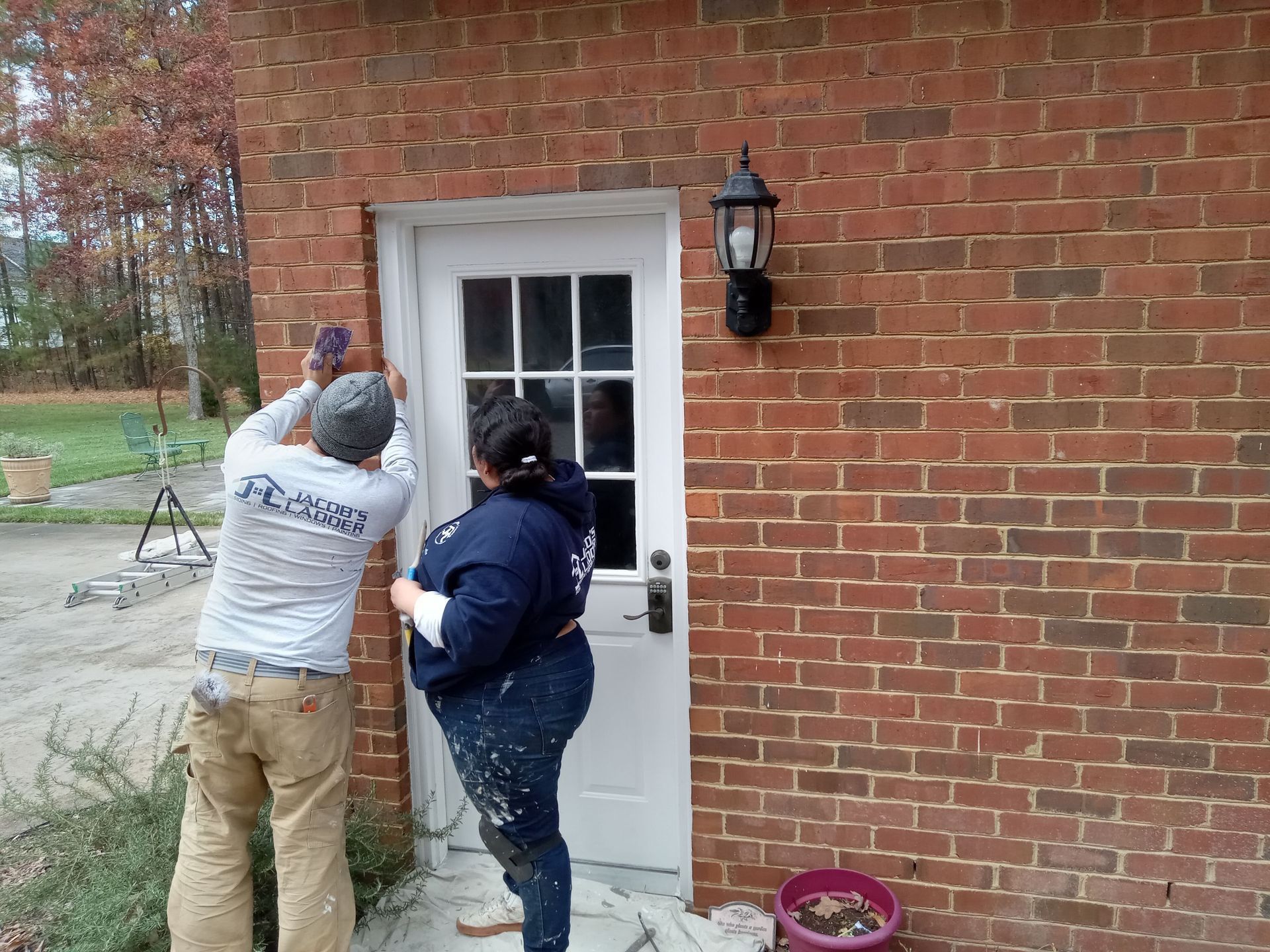 Two people painting a white door on a brick building's exterior. One sands, the other applies paint.
