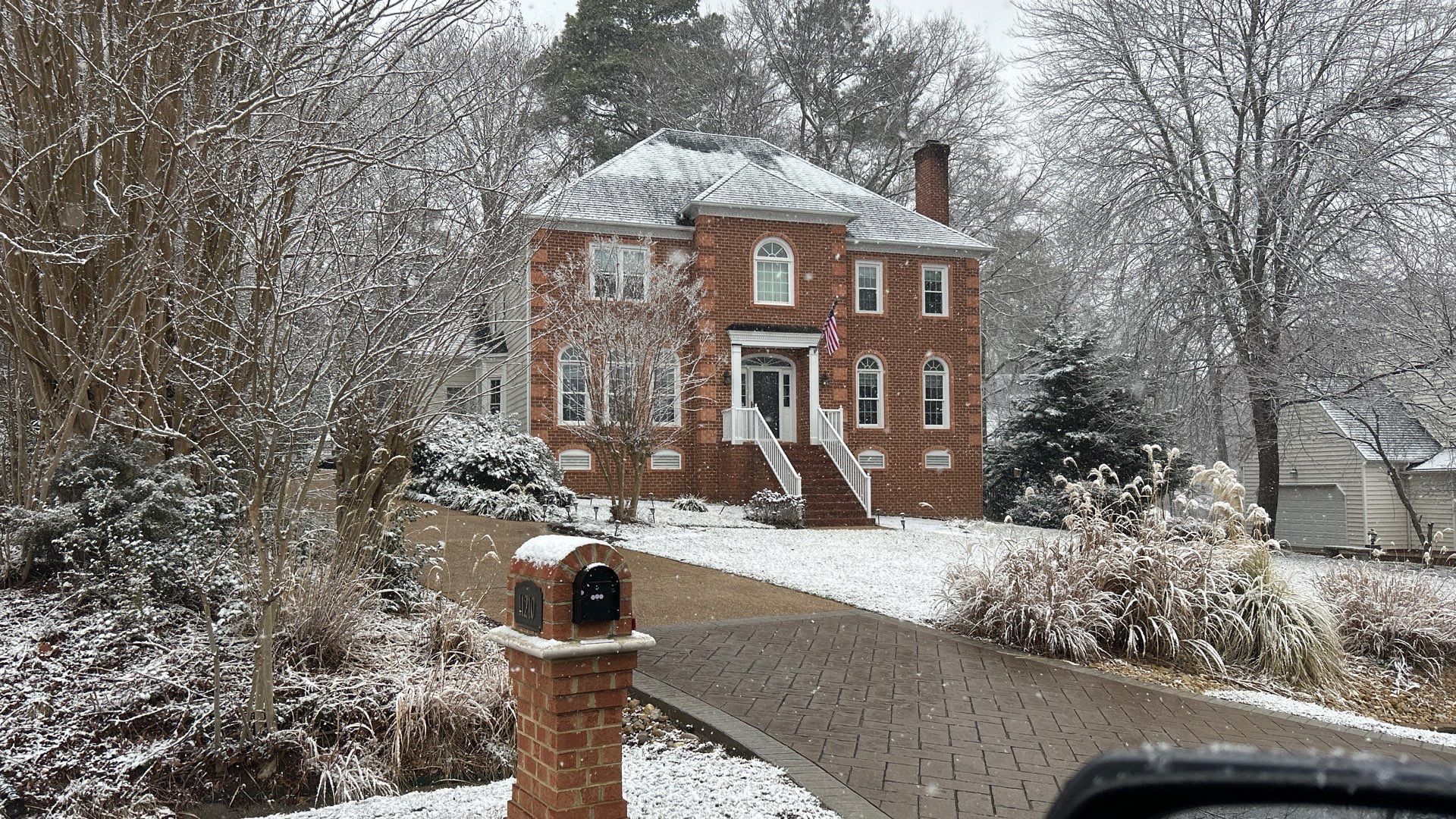 A brick house with snow-covered roof and surroundings, snow falling on the driveway.