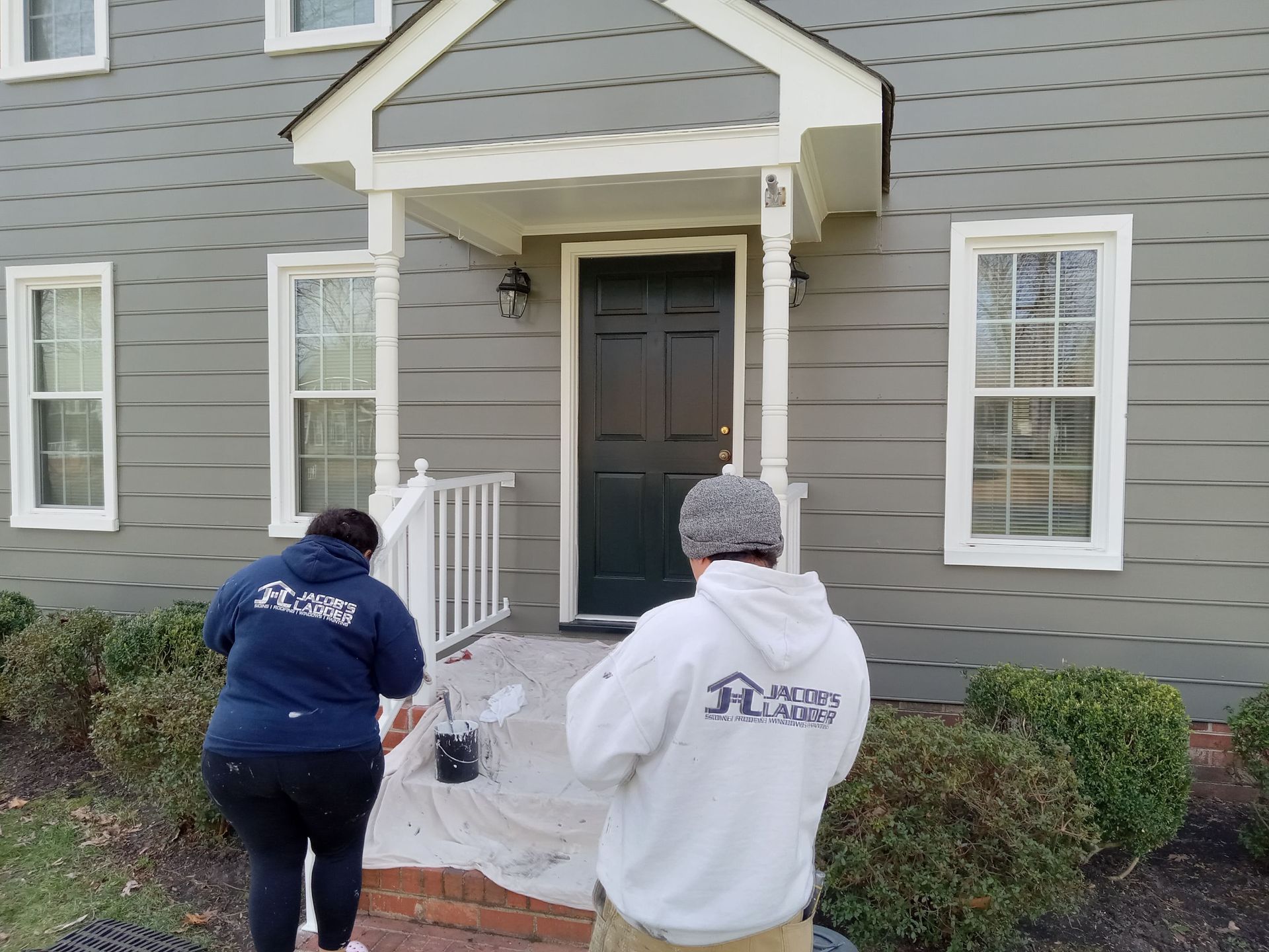 Two people painting a house's front porch. The house has gray siding and white trim.