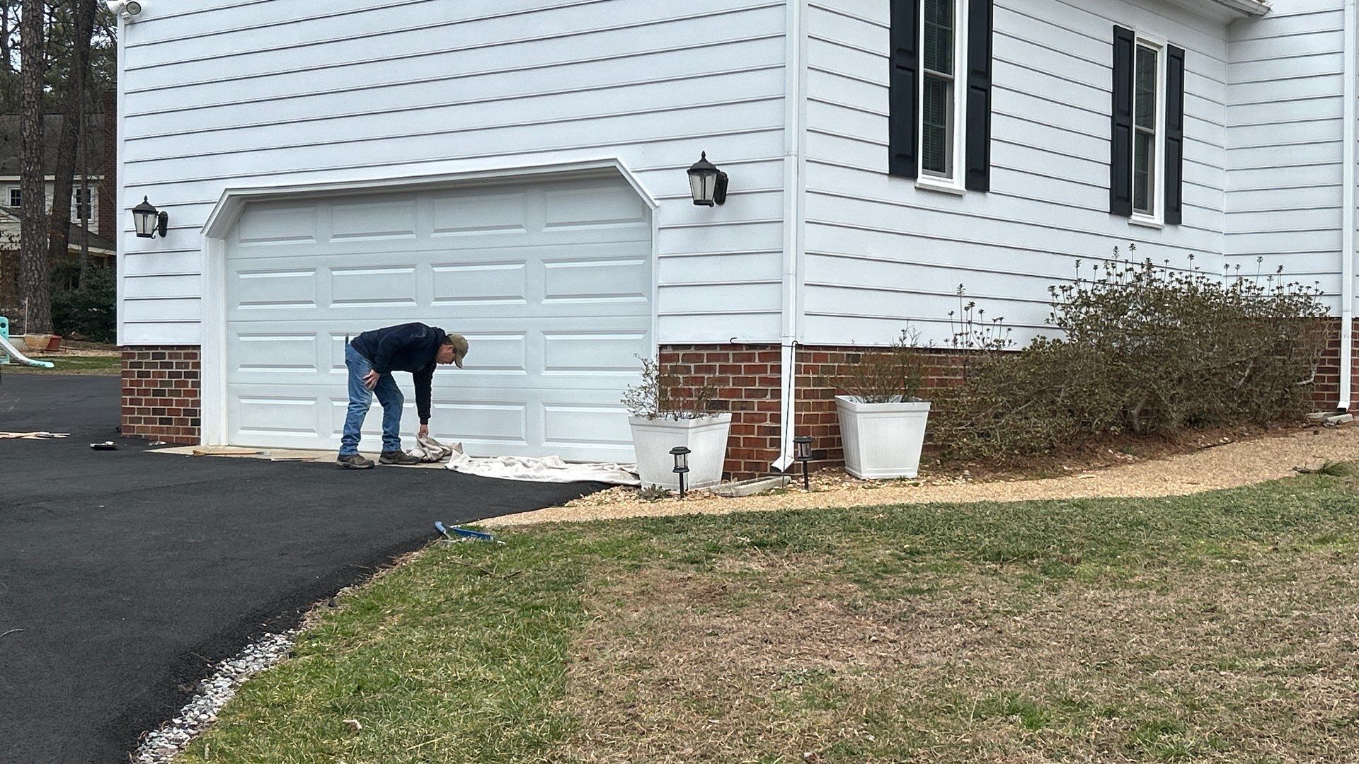 Person near a white garage door, working on the ground with tools, in front of a house. Asphalt driveway and lawn visible.