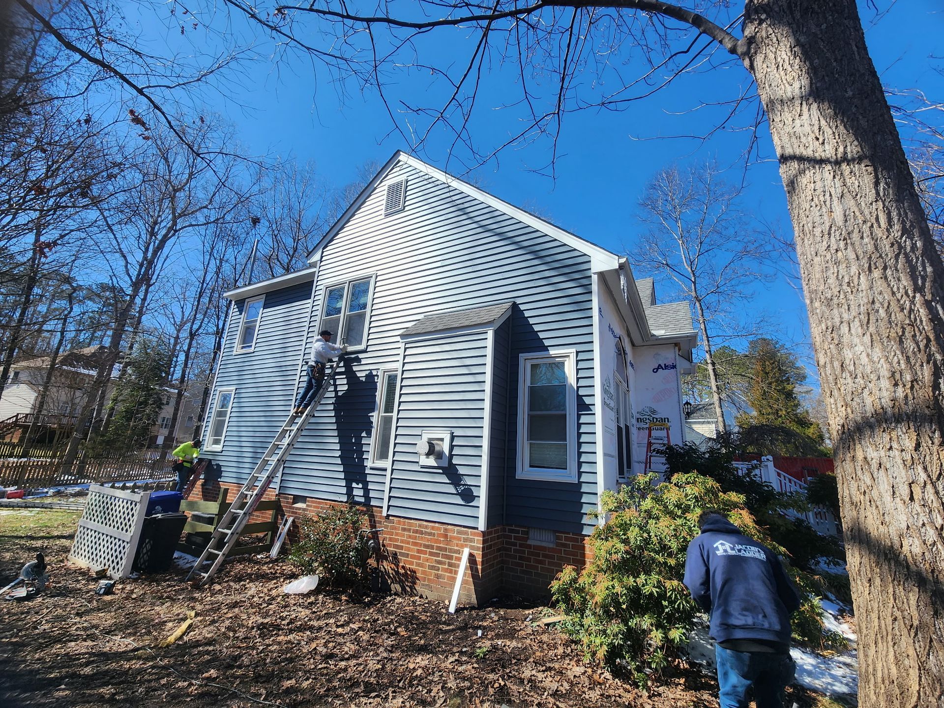 House with blue siding under construction, two workers on ladders, sunny day.