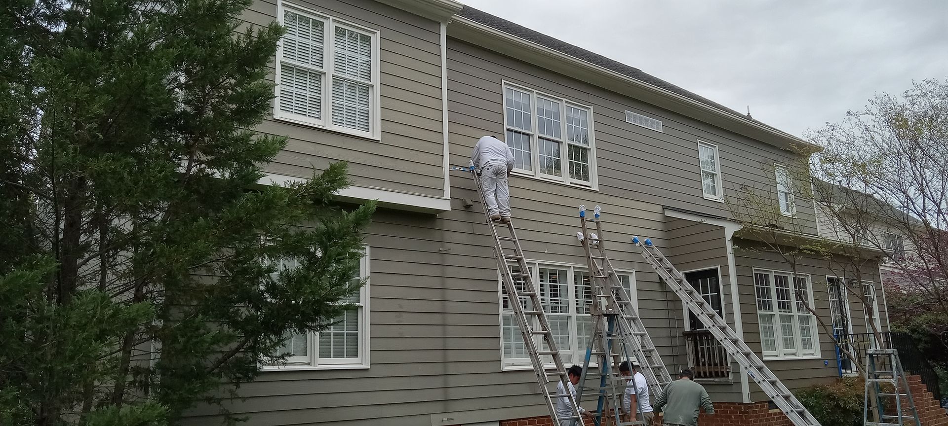 Workers on ladders painting the exterior of a two-story house. Gray siding, white trim, cloudy sky.