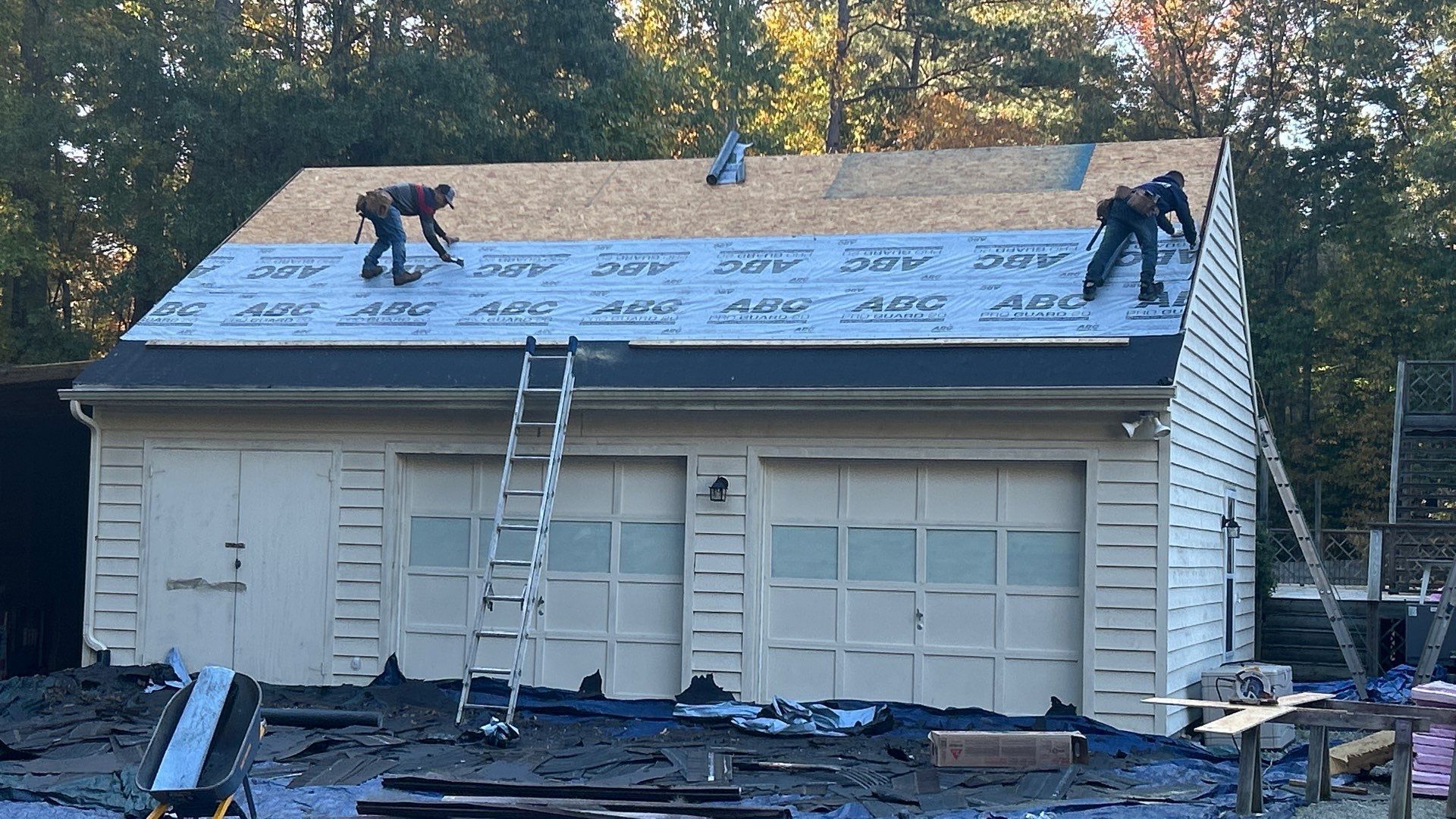 Roofers working on a garage roof, laying roofing material. Ladder and tools visible.