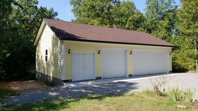 Yellow three-bay garage with white doors, brown roof, and gravel driveway, set against trees.