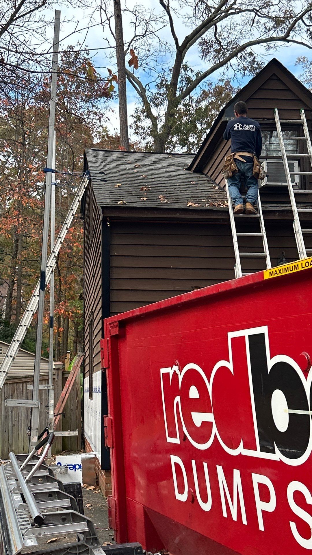 Man on ladder near brown house with a red dumpster in the foreground.