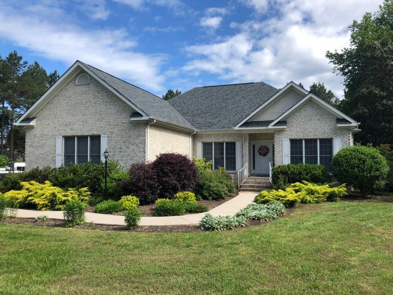 Light brick house with gray roof, surrounded by green lawn and colorful landscaping.