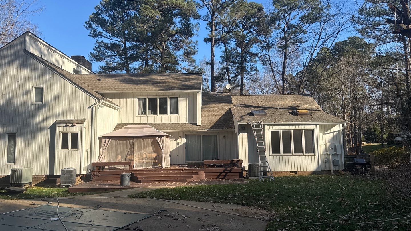 Back of a two-story white house with a partially replaced roof, a wooden deck, and tall trees.