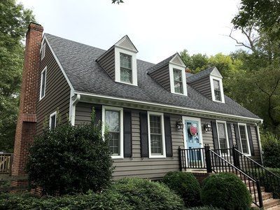 Tan-sided house with dark roof, brick chimney, and black shutters, with three dormer windows and a blue front door.