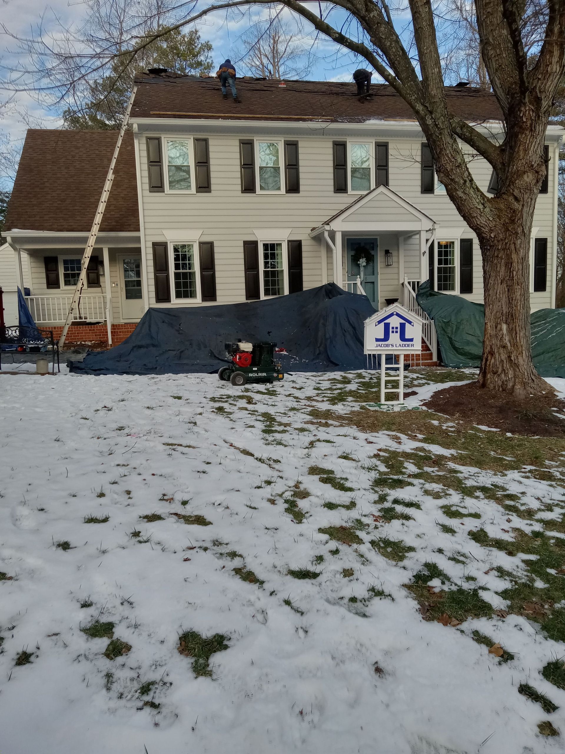 Roofers working on a house with snow on the ground. Tarps cover the lawn, ladder leaning on the house.