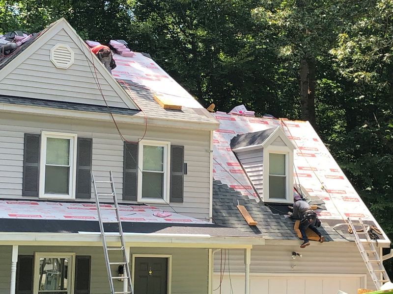 House roof being worked on; worker installs shingles, pink underlayment visible. Gray siding, two-story.