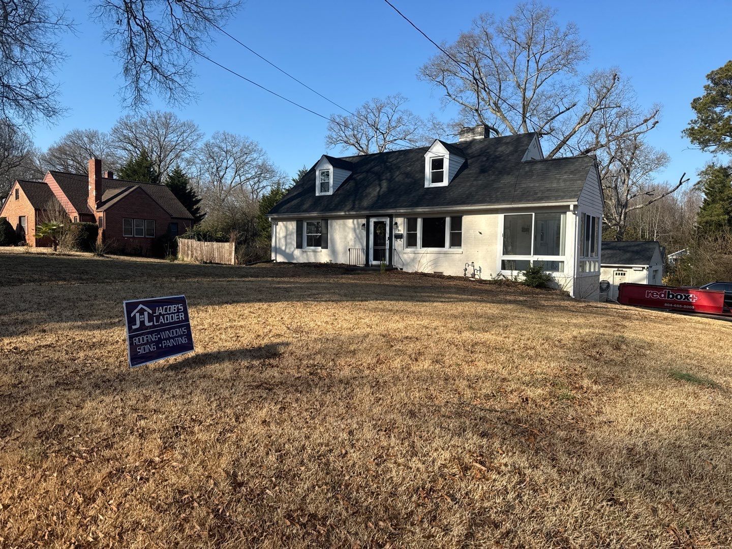A house with a sign in front, brown grass, and trees under a clear blue sky.
