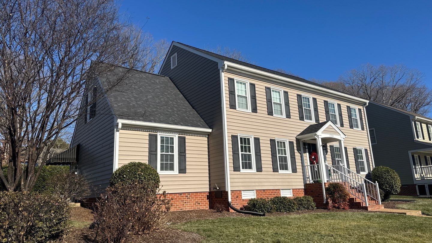 Two-story house with tan siding, black shutters, brick base, and a dark roof under a blue sky.