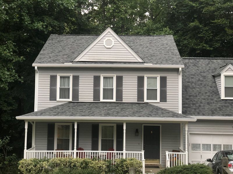 Two-story gray house with dark gray shutters, white trim, and a porch, set against a green tree background.