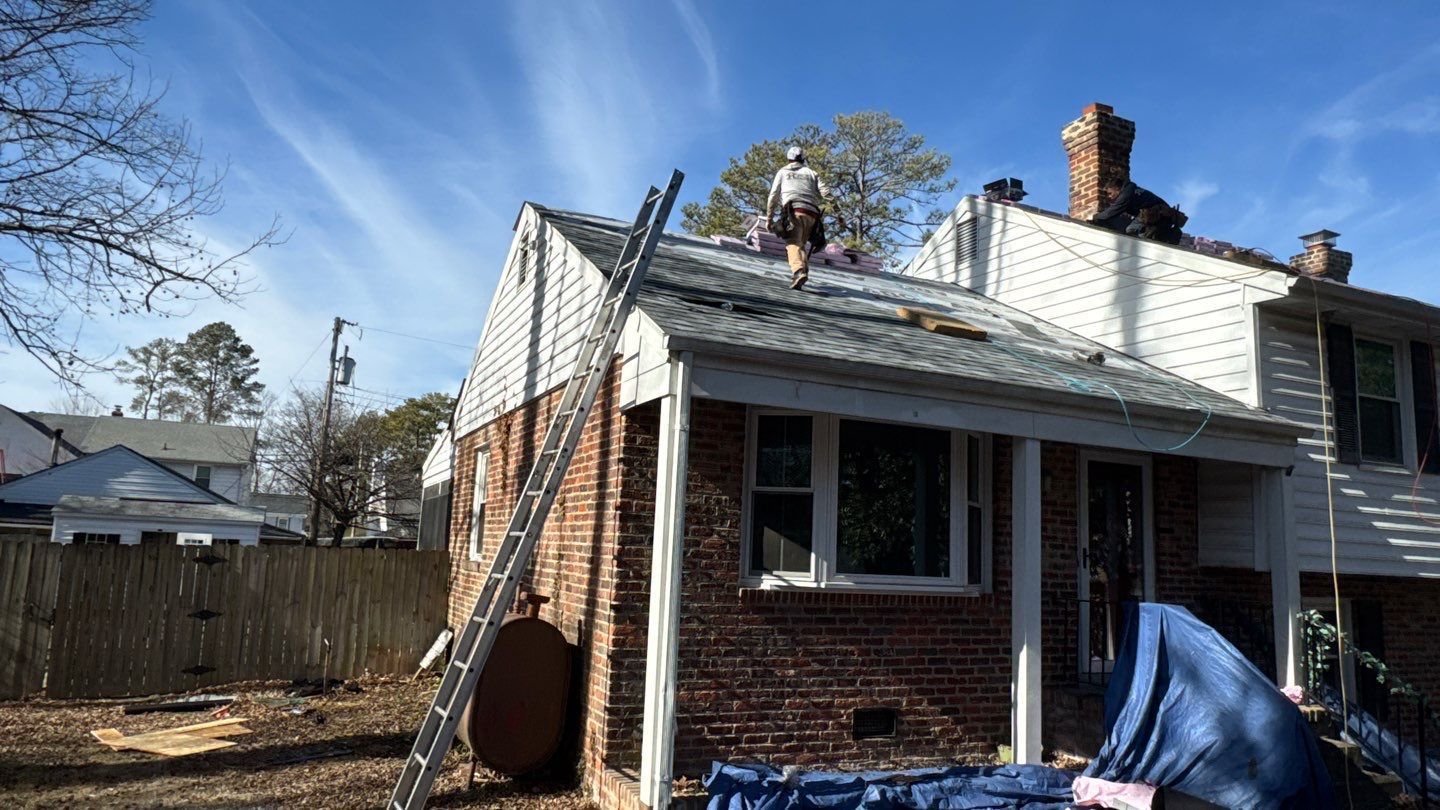 A person on a rooftop repairs shingles. A ladder leans against the roof; blue tarp covers something on the ground.