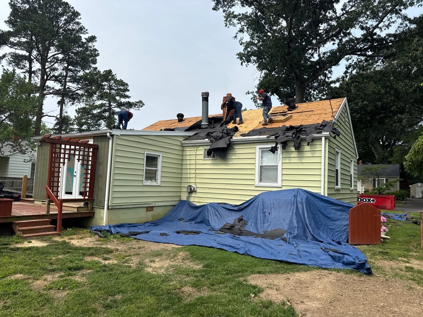 Roofing crew working on removing old shingles from a light green house. A blue tarp protects the ground.