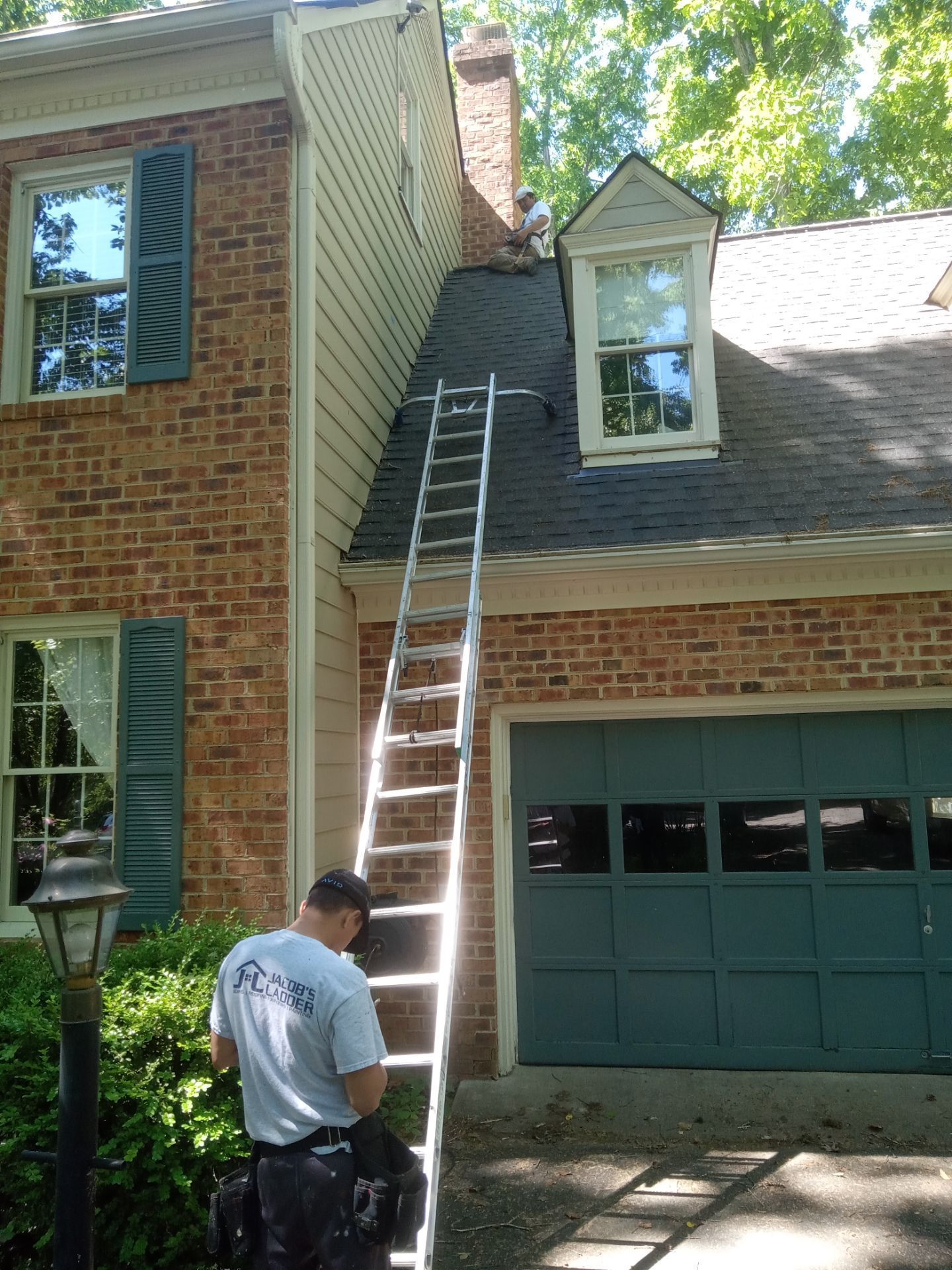 A man on a ladder near a chimney on a two-story brick house with a green garage door and shutters.