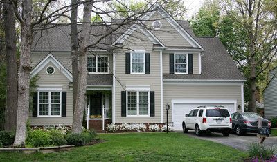 Two-story beige house with a garage; white SUV and gray car in the driveway. Trees and green grass in front.