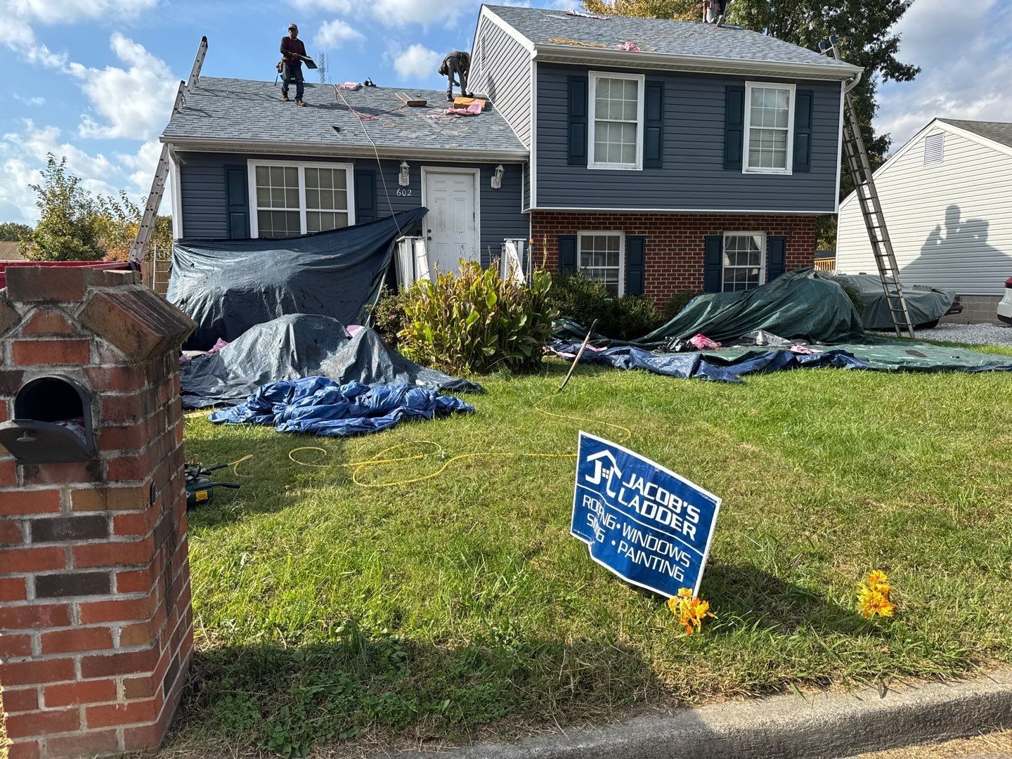 Roofers working on a house with blue siding. Tarps cover the lawn. A sign for a roofing company is in the yard.
