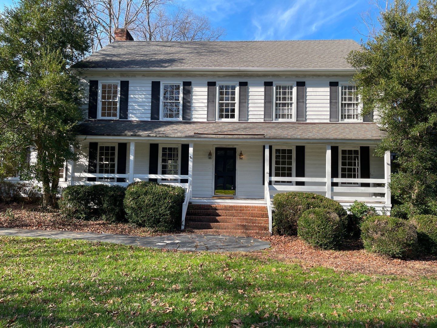 Two-story white house with black shutters, front porch, and red brick steps, surrounded by green shrubs and trees.