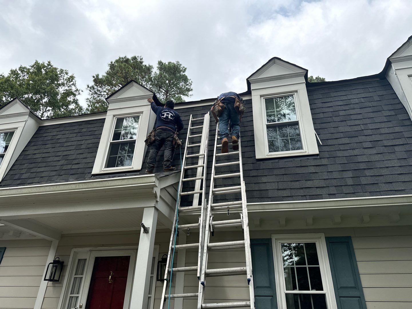 Two people on ladders working on a black shingle roof. White dormer windows and cloudy sky visible.