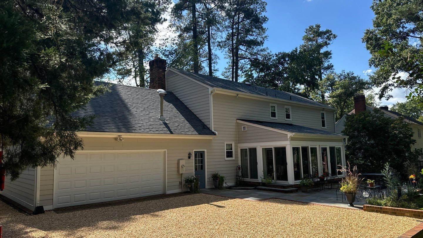Beige house with a two-car garage, a sunroom, and a gravel driveway, surrounded by trees.