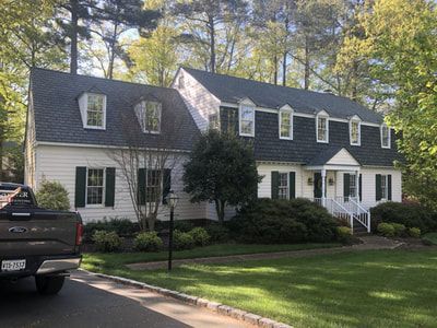 White two-story house with green shutters, dormer windows, and a dark roof; a pickup truck is parked in the driveway.