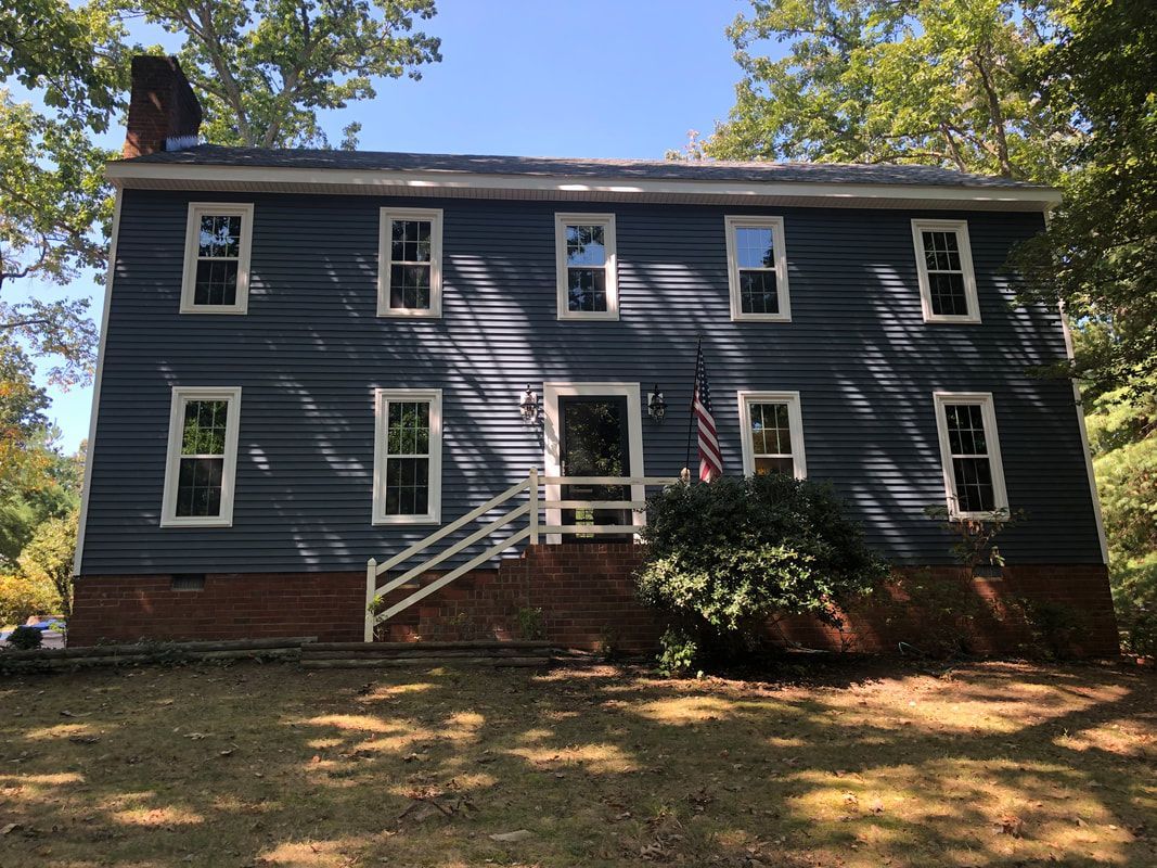 Two-story blue house with white trim, brick foundation, front door, and an American flag.