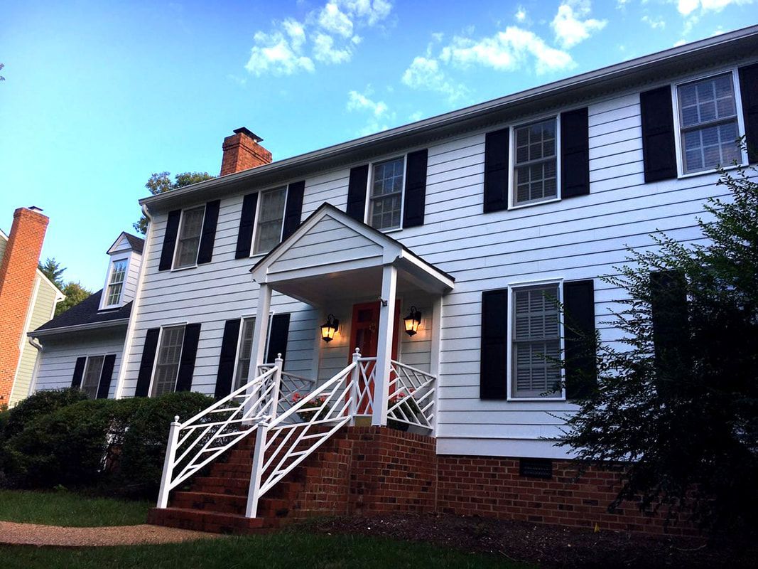 White two-story house with black shutters, red brick foundation, and a small front porch under a partly cloudy sky.