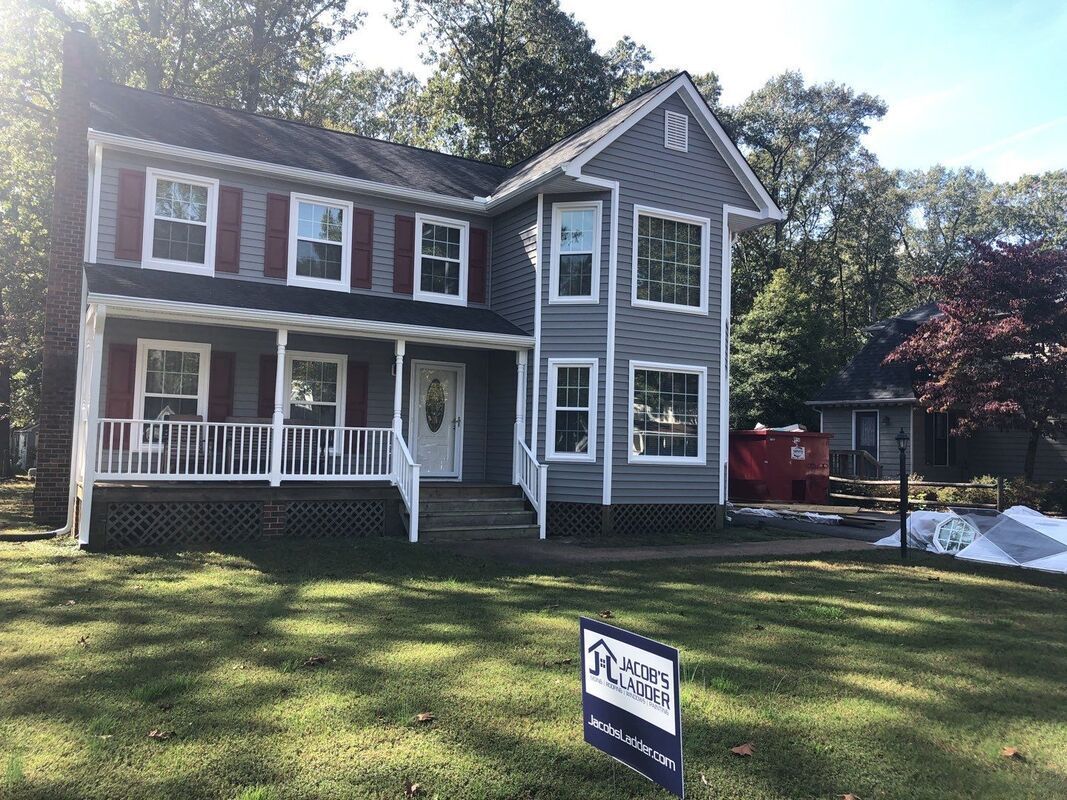 Two-story house with gray siding, red shutters, and a porch, on a grassy lawn with a real estate sign.