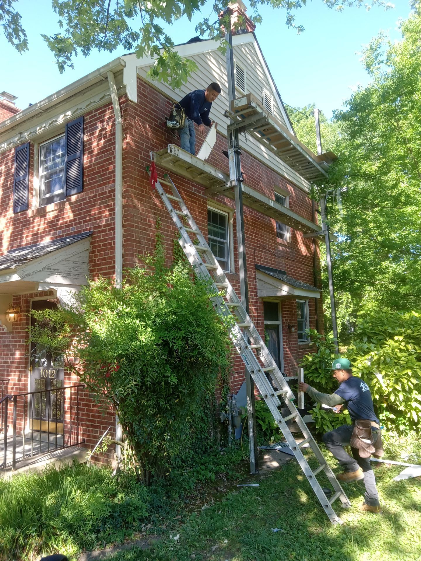 Two workers on scaffolding repair a brick house's trim. One worker hands materials to the other.