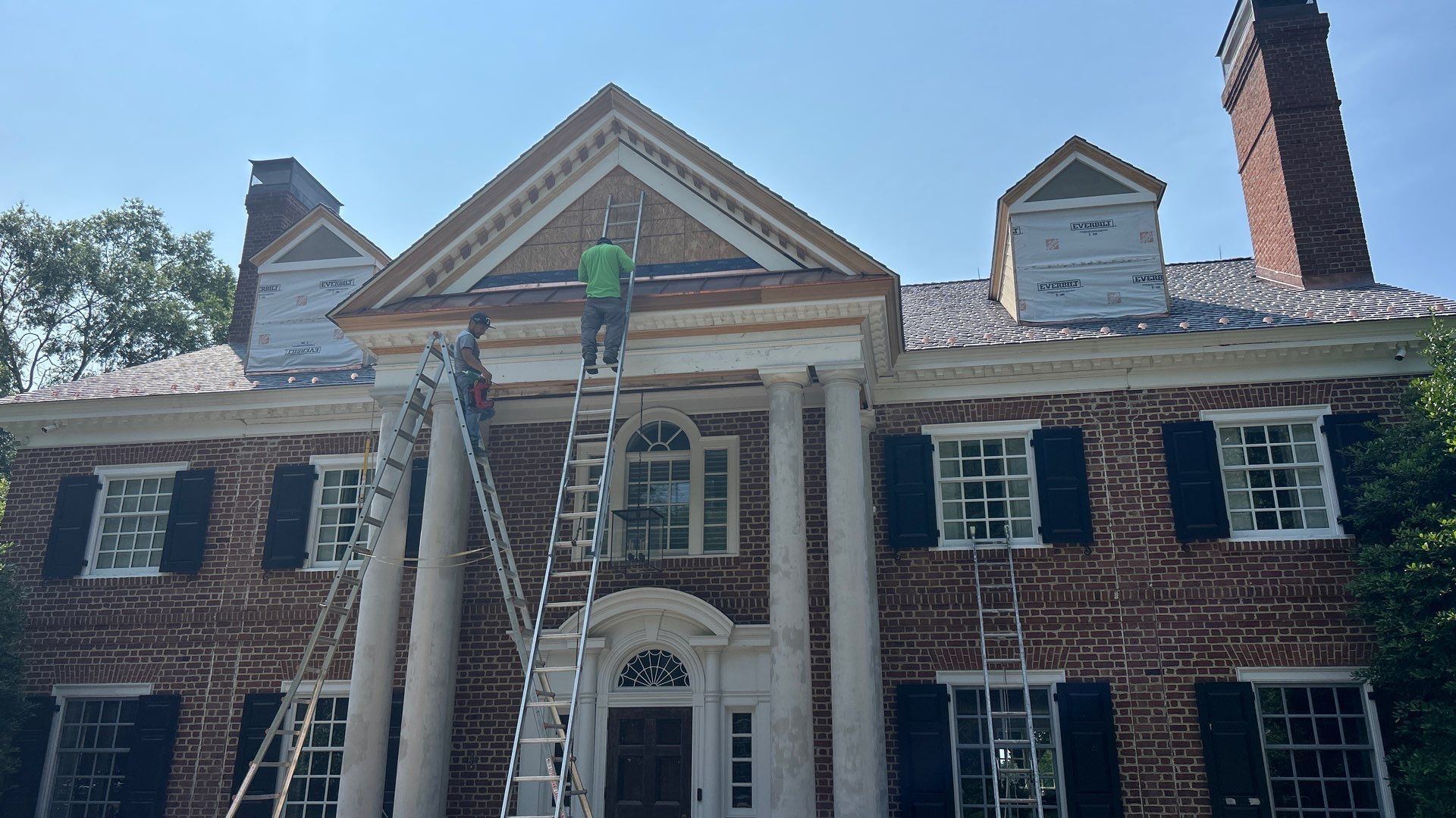 Man on a ladder repairing a brick house with white columns, under a blue sky.